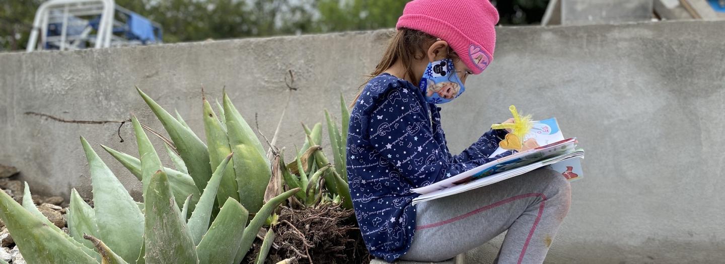 Mädchen sitzt mit Maske und pinker Mütze neben Aloe-Vera Pflanzen