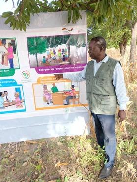 Ein Mann, der auf einem Plakat, dass unter einem Baum aufgespannt ist, etwas zeigt.