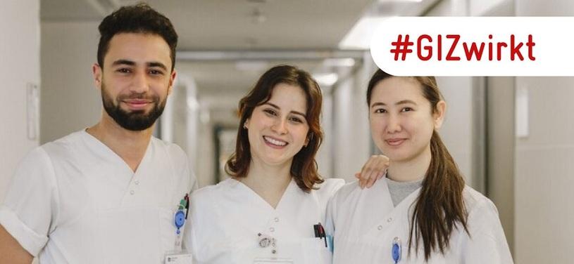 Drei Pflegefachkräfte stehen in einem Krankenhausflur und lächeln in die Kamera.