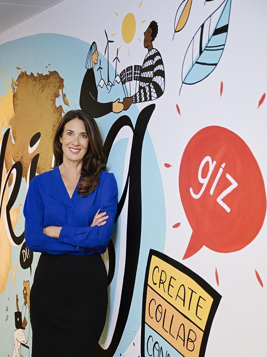 Eine Frau mit langen braunen Haaren in einer blauen Bluse steht mit verschränkten Armen vor einer bunten Wand mit der Aufschrift „giz“ und Illustrationen zu Nachhaltigkeit und Zusammenarbeit.