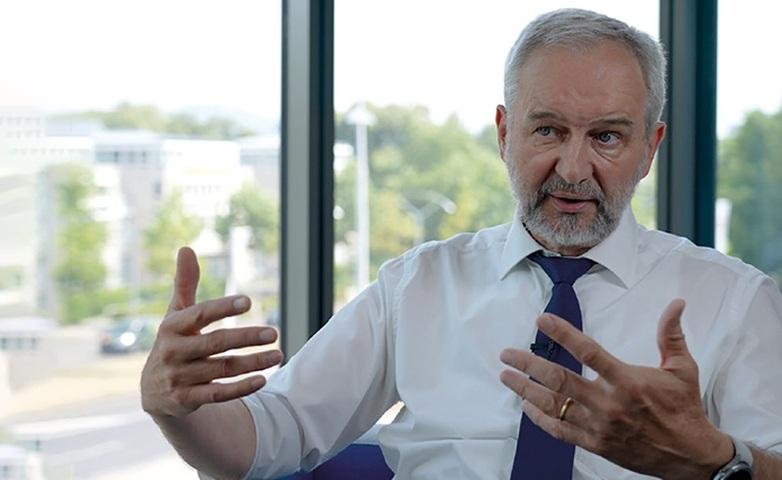 Bild von Albert Engel: Ein grauhaariger Mann in weißem Hemd nit dunkelblauer Krawatte und Bart sitzt vor einem Fenster mit grüner Hintergrundkulisse und hat die Hände gestikulierend erhoben.