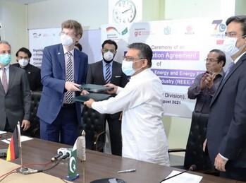 Zwei Männer mit Mundschutz übergeben einander Dokumente. Neben und hinter ihnen stehen sechs weitere Männer, die teilweise applaudieren.