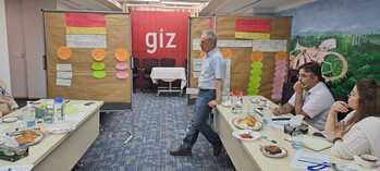 Menschen sitzen und stehen bei einer strategischen Planungssitzung in einem Raum zusammen. Im Hintergrund stehen Schreibwände mit Notizen.