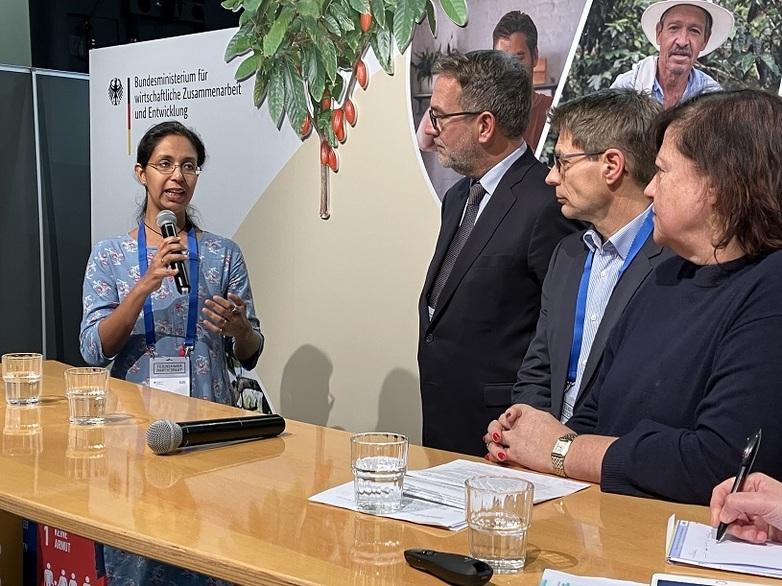 Eine Frau mit Mikrofon steht vor einem Tisch und spricht mit drei weiteren Personen.