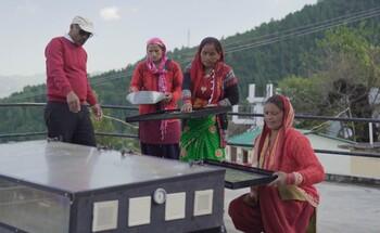Eine Gruppe von Frauen und ein Mann arbeiten in einer ländlichen Umgebung auf einem Dach an einem solarbetriebenen Lebensmitteltrockner und zeigen umweltfreundliche Konservierungstechniken.