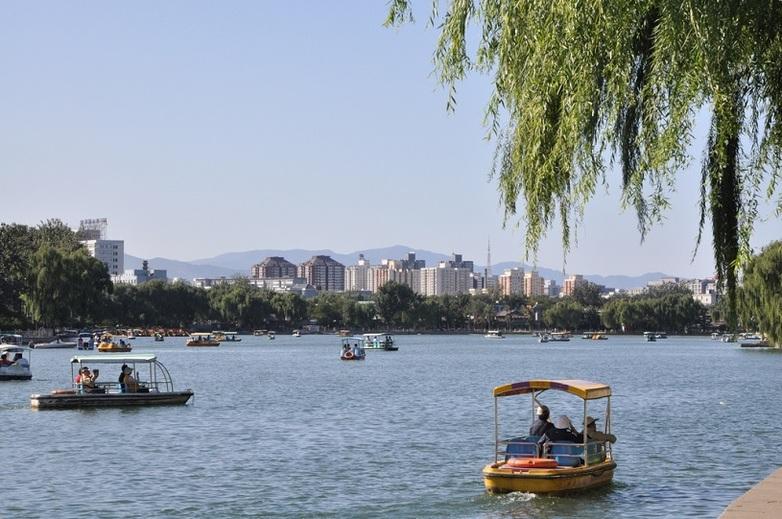 Blick über einen Fluss in Bejing. Es fahren viele kleine Boote auf dem Fluss. Am Horizont sieht man mehrere Wohngebäude.