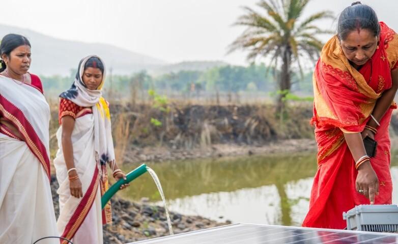Frauen bedienen eine solarbetriebene Wasserpumpe in der Nähe eines Teiches in einer ländlichen Gegend in Indien.