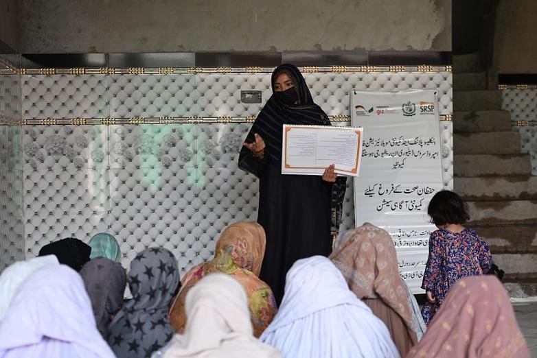 A woman holding a document in one hand and making a gesture with the other. A group of women are sitting in front of her listening. Behind her is a display which includes the GIZ logo.