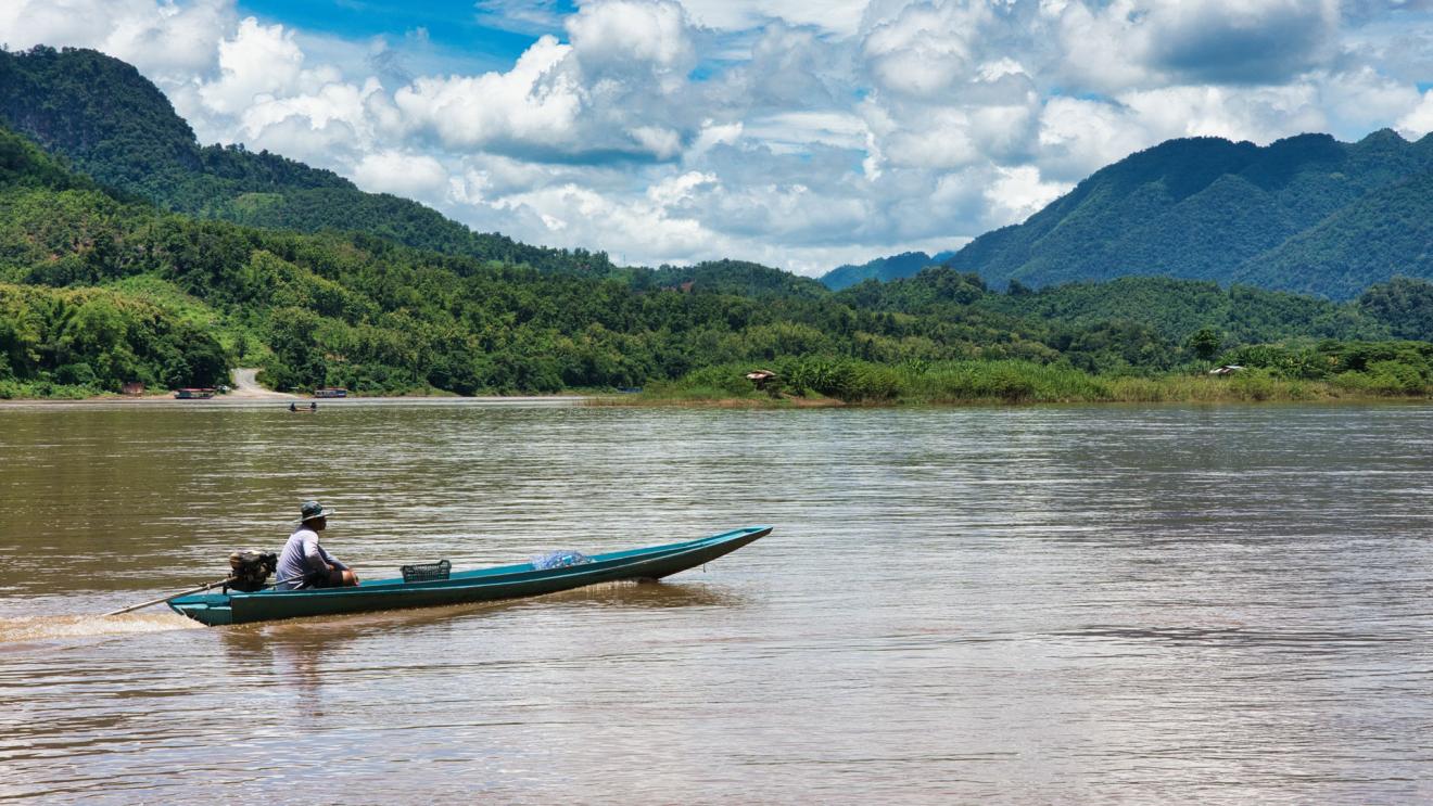 Ein Mann fährt mit seinem Boot auf dem Mekong. Im Hintergrund bewaldete Berge.