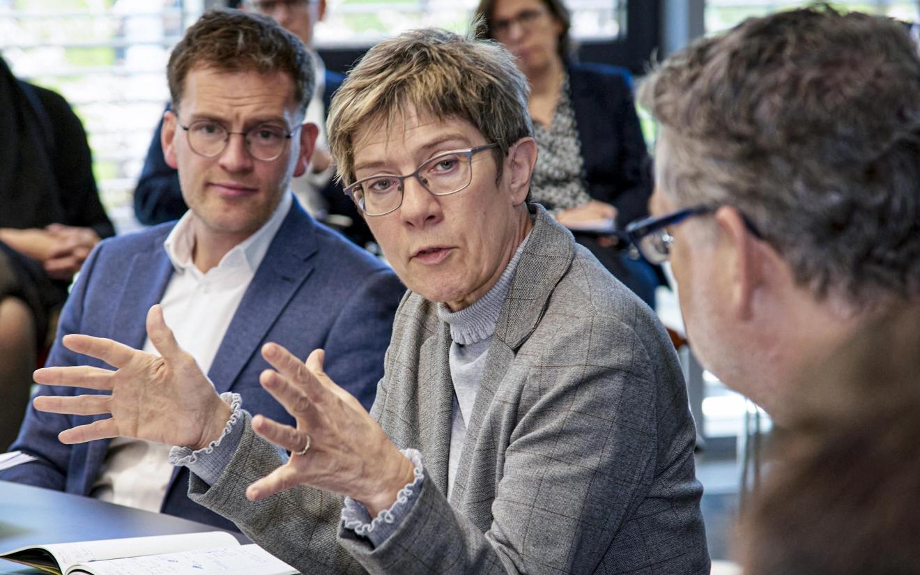 Diskussion in einem Konferenzraum mit einer Frau im grauen Blazer, die mit Gestik spricht, umgeben von mehreren Personen in Businesskleidung, die aufmerksam zuhören.