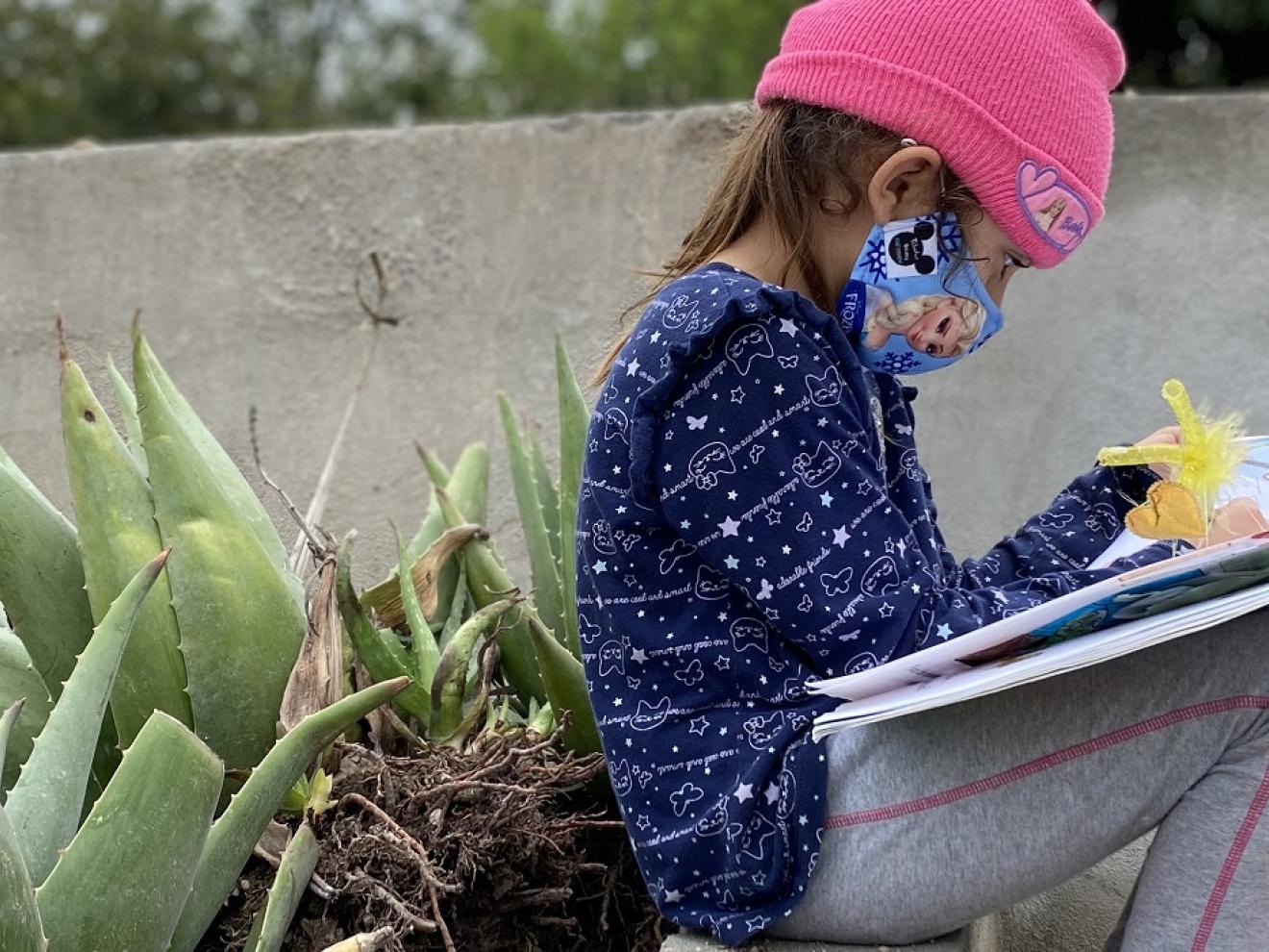 Mädchen sitzt mit Maske und pinker Mütze neben Aloe-Vera Pflanzen