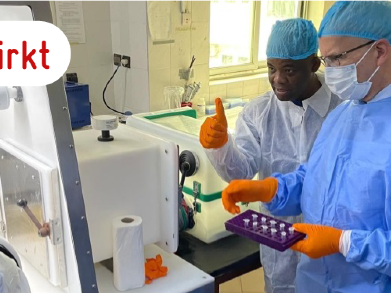 Fünf Personen in medizinischer Schutzkleidung und Maske stehen um ein Laborgerät herum.