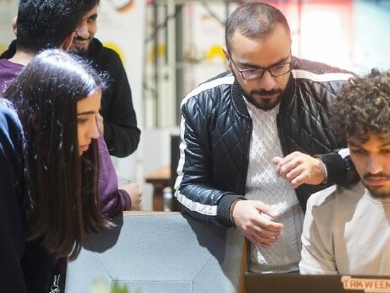 Fünf junge Menschen in einem modernen Raum, drei davon blicken auf ein Laptop, zwei unterhalten sich.