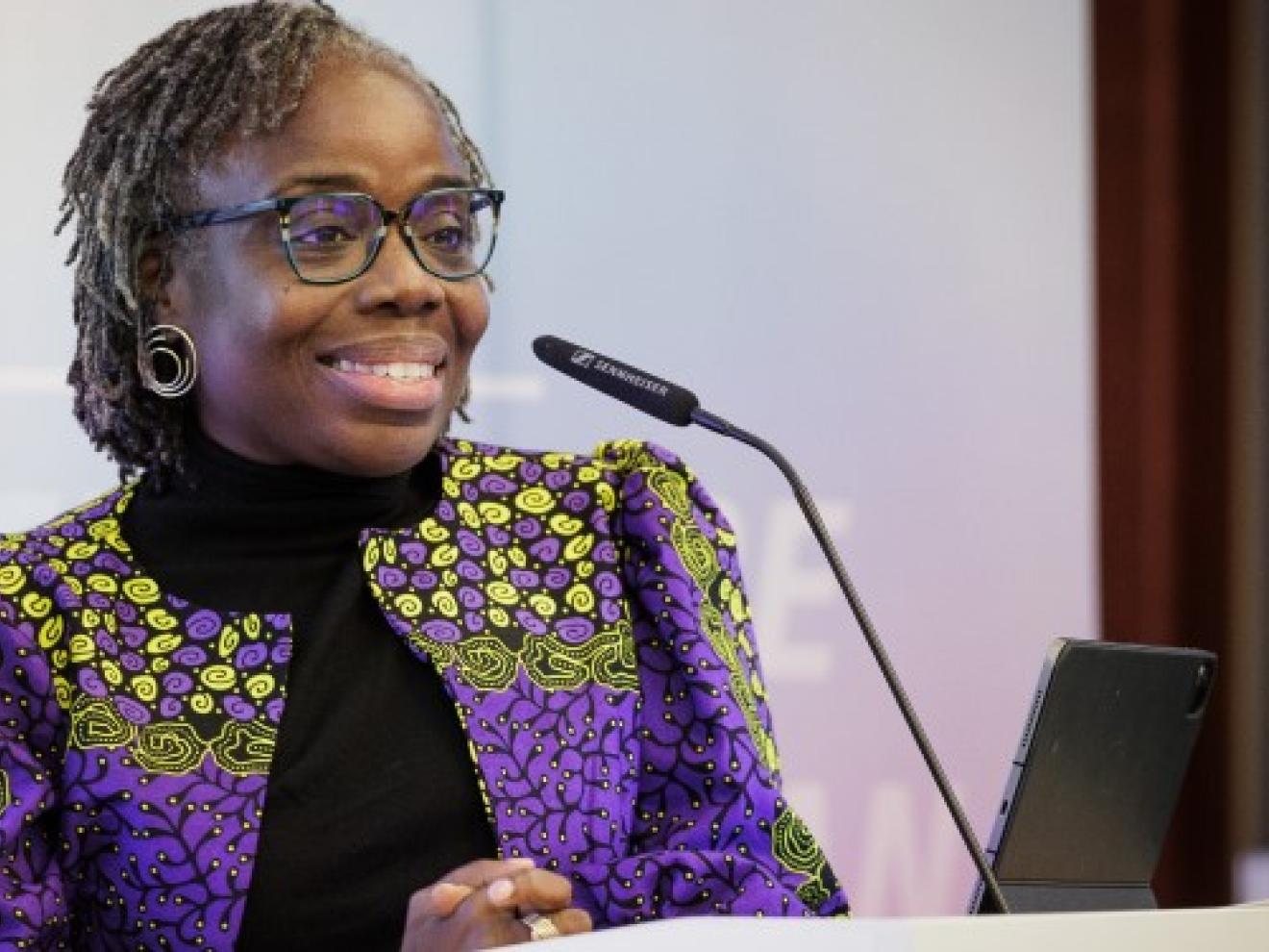 Eine Frau (Mavis Owusu-Gyamfi) in einem lila Blazer steht an einem Podium mit einem Mikrofon und lächelt.