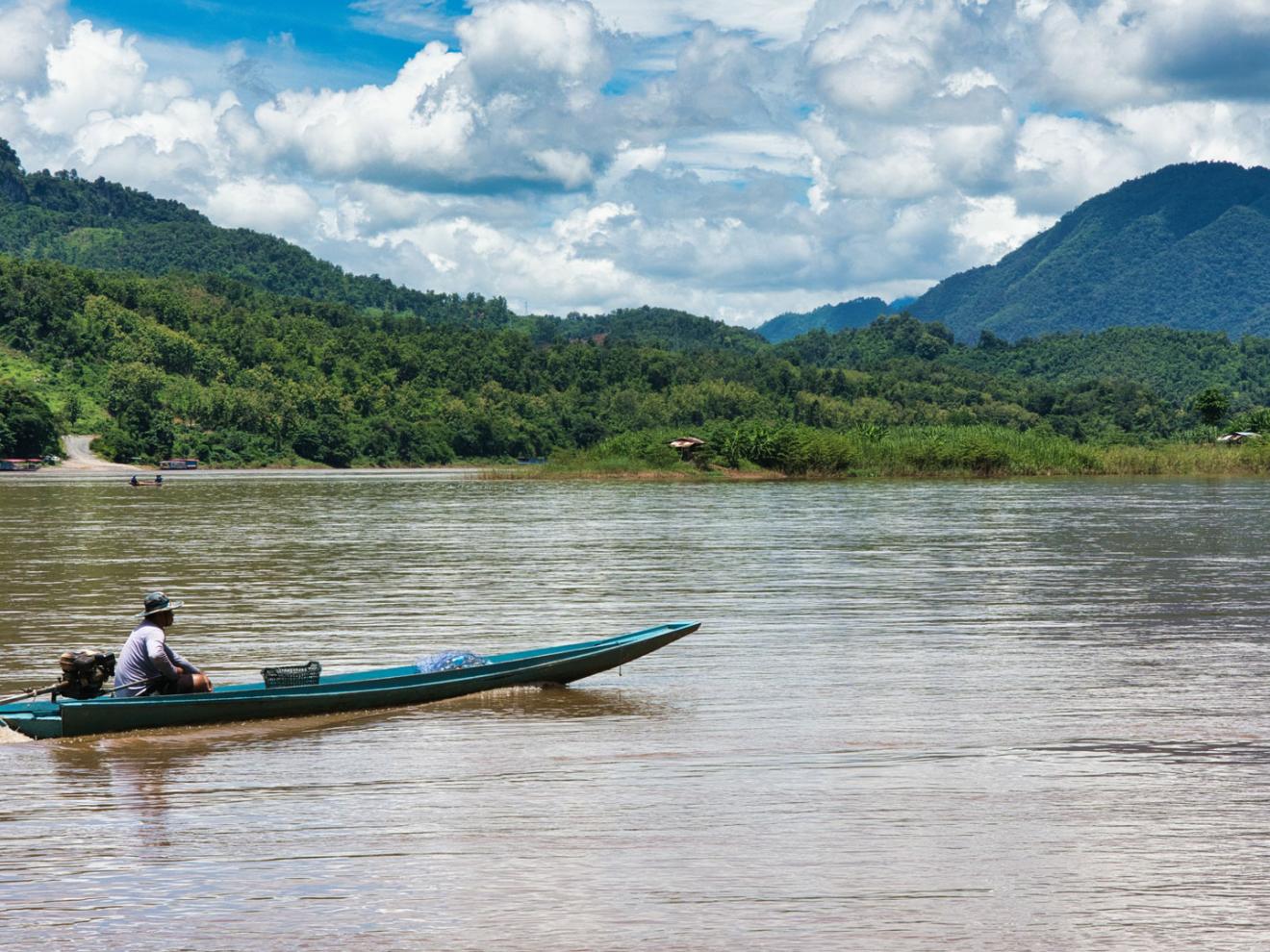 Ein Mann fährt mit seinem Boot auf dem Mekong. Im Hintergrund bewaldete Berge.