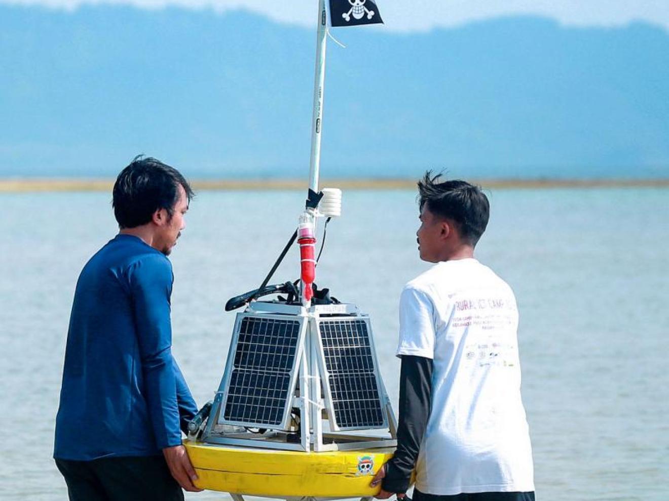 Zwei Männer tragen eine solarbetriebene Messboje durch flaches Wasser, im Hintergrund sind Berge und eine Sandbank zu sehen.