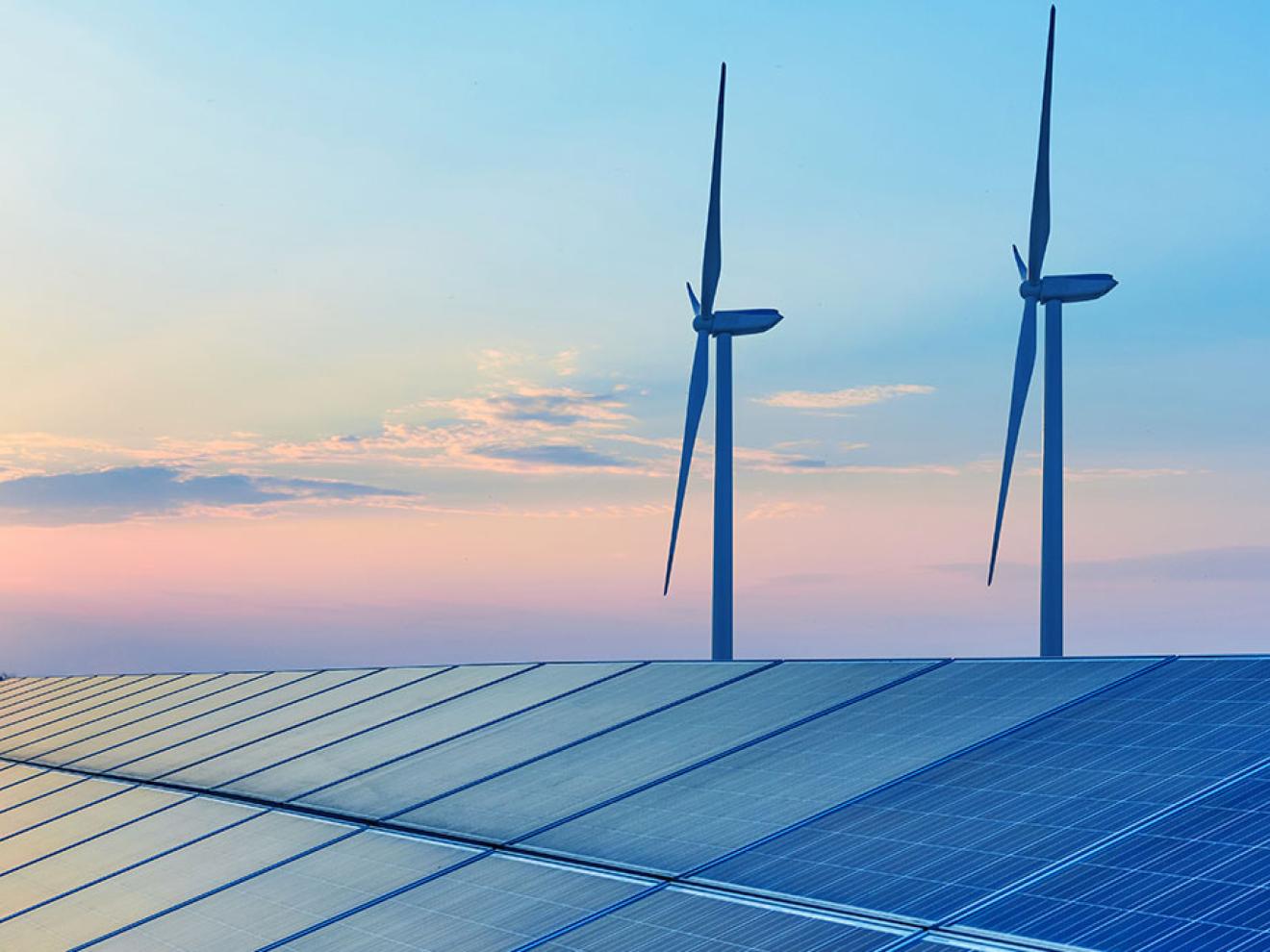 Eine Reihe von Solaranlagen im Vordergrund mit drei Windrädern dahinter, abgebildet vor einem Himmel in blauen und rosafarbenen Abendtönen.