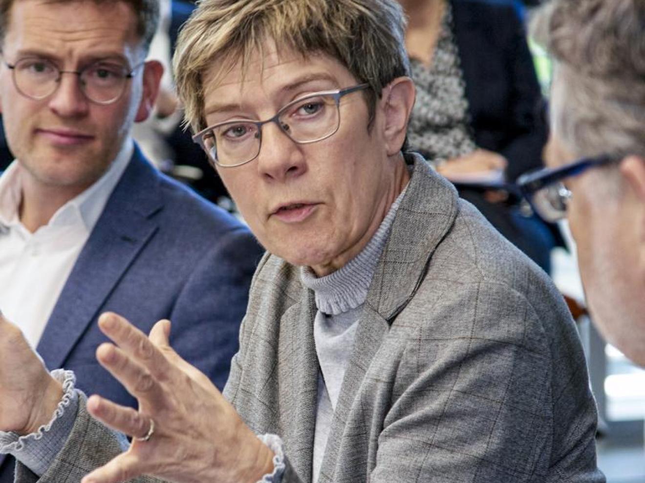 Diskussion in einem Konferenzraum mit einer Frau im grauen Blazer, die mit Gestik spricht, umgeben von mehreren Personen in Businesskleidung, die aufmerksam zuhören.