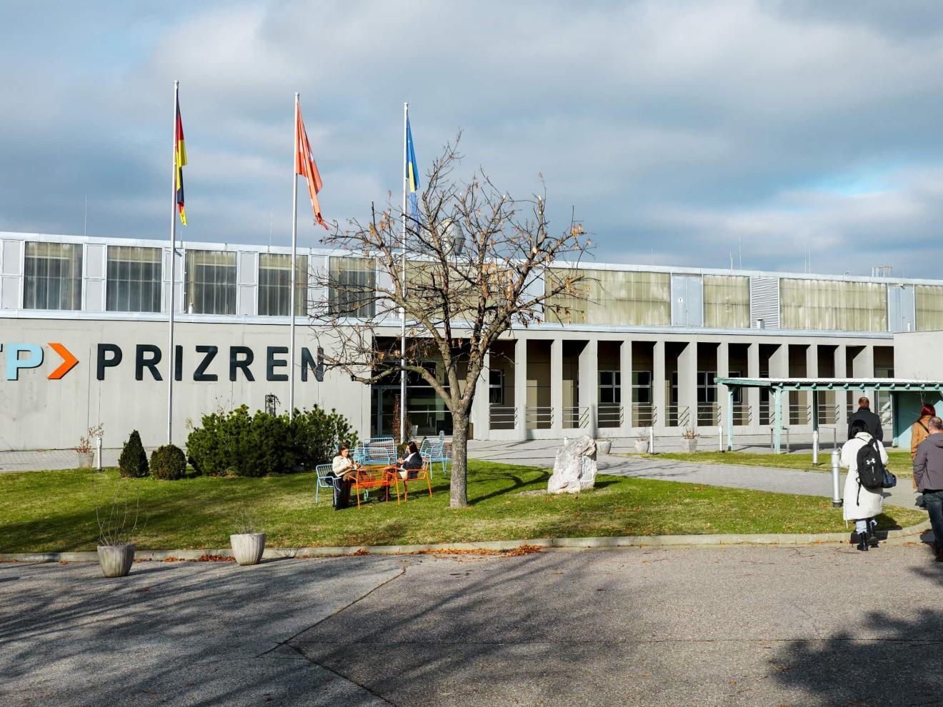 Außenansicht des ITP Prizren-Gebäudes mit drei Fahnenmasten, Sitzgelegenheiten auf der Wiese und mehreren Personen auf dem Gelände.