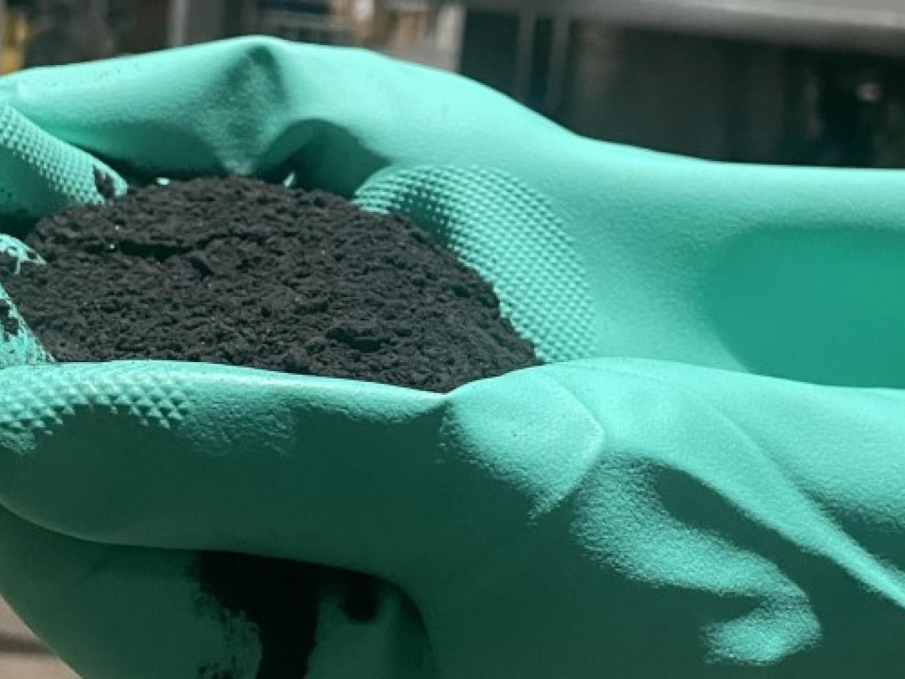 Hände in grünen Gummihandschuhen, die ein schwarzes Pulver halten.