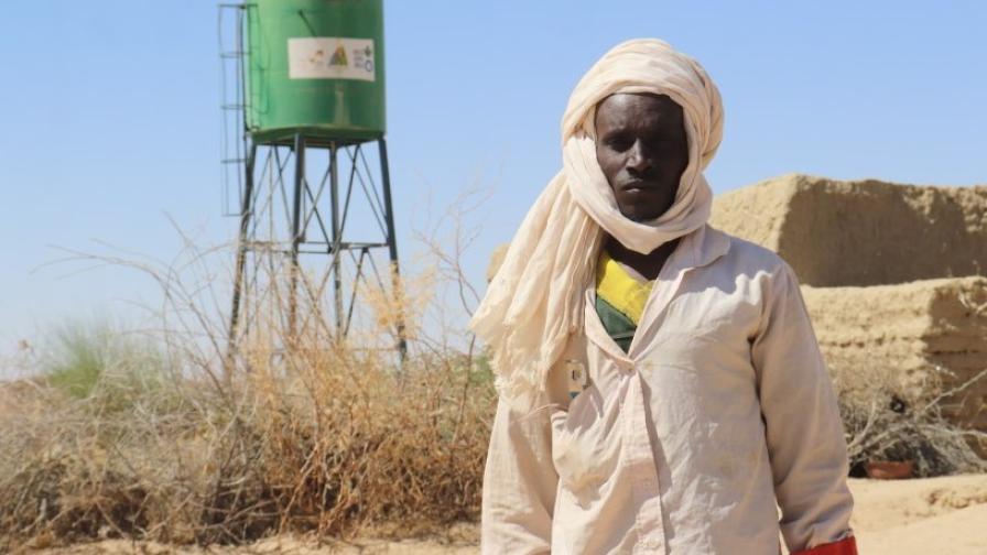 Ein Mann – Ibrahim Yatara – steht in Wüstenkleidung vor einem grünen Wassertank in einer trockenen Landschaft.