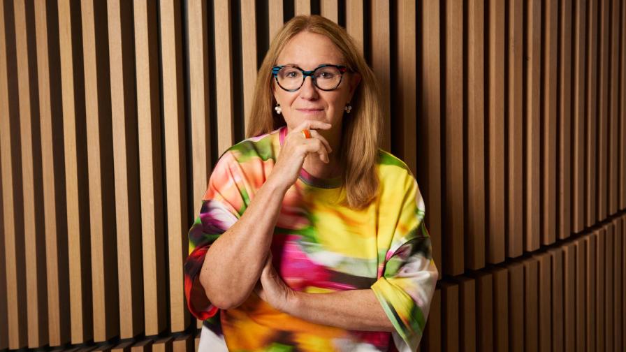 Eine Frau mit langen blonden Haaren und Brille steht vor einer Wand mit Holzlamellen und trägt eine bunte Bluse.