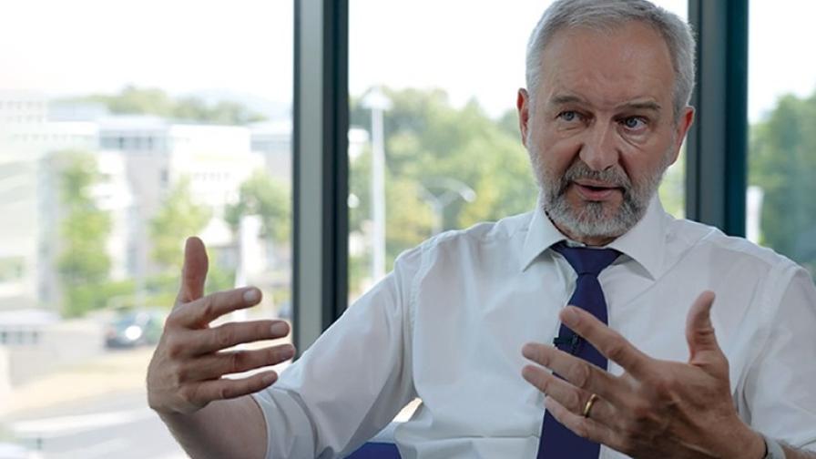 Bild von Albert Engel: Ein grauhaariger Mann in weißem Hemd nit dunkelblauer Krawatte und Bart sitzt vor einem Fenster mit grüner Hintergrundkulisse und hat die Hände gestikulierend erhoben.